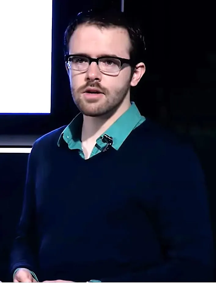 Brian Christian speaking at a TEDx event, in glasses and a dark sweater